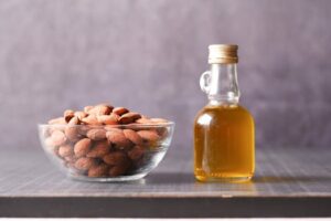 A close-up of almonds in a glass bowl alongside a bottle of almond oil.