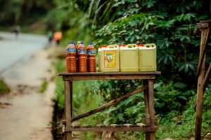 Rural roadside stall featuring bottles and jerrycans of oil and juice with a lush green backdrop.